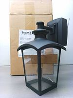 The black outdoor wall lantern in front of its shipping box showing the lantern's shape, glass, and mount.