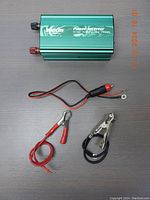Green 3000W car power inverter lying flat with two power cables, one with alligator clamps and one with cigarette lighter and ring terminals.