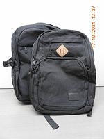 Two black backpacks side by side showing front views, different design details visible.