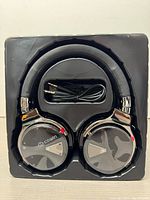 Front view of Cowin E7 headphones in black plastic tray showing outer earcup design, foldable headband, and included audio cable.