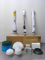 Electric spin scrubber with 3 detachable handle parts displayed standing on box alongside accessories including mounting brackets and suction base.