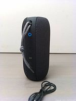 Front view of Monster S320 speaker standing upright, showing fabric mesh surface, carry cord looped and rubberized control buttons lit with blue light.