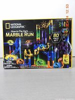 Front of box showing National Geographic Glow-In-The-Dark Marble Run branding, colorful images of translucent plastic track pieces in yellow, blue, and purple, glowing galaxy-patterned marbles, and '80 Piece Set' text.