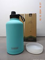 Single blue KOODEE insulated water bottle with black lid, white silicone base, and packaging box in background. Clear brand marking on bottle.