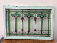 Front view of stained glass window showing full rectangular design framed in painted wood with hanging hooks on top.