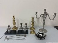 Full lot view showing all candlesticks, candelabra, black metal candle holder, candle snuffer, thimble, and floral candle holder arranged on table.