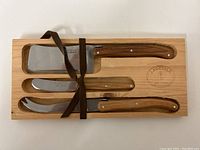 Full view of wooden box containing three Laguiole cheese cutting utensils tied with a dark brown ribbon.