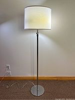 Full view of the floor lamp showing the chrome base, pole, and white fabric lampshade, lit from inside.