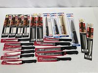 Photo showing multiple kitchen knives and steak knives in original packaging from Good Cook and Diamond Cut. Includes steak knives, carving knives, and slicing knives with black handles lined up in two rows.