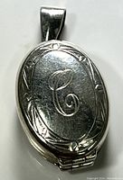 Close-up front view of an oval sterling silver locket with engraved monogram letter 'C' and decorative border.