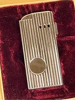 Close-up image of silver-tone metal lighter with vertical ridges and circular accent, placed inside velvet lining of box.