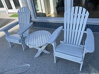 Two white Adirondack chairs and the small round side table positioned outside on a concrete surface.