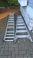 Two aluminum ladders side by side on a stone paved surface by a stone wall and house. One is an extension ladder, the other a step ladder.