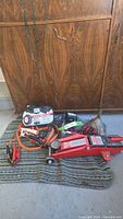 Wide shot of all items in the lot including trolley jack, jumper cables, trickle chargers, and tire chains on floor in front of wooden cabinet.