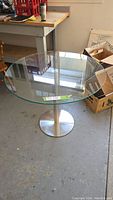 Full view of the round clear glass kitchen table showing the glass top and stainless steel pedestal base.