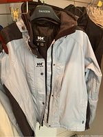 Front view of the Helly Hansen men's jacket showing light blue and black color scheme, zipper, and buttons.