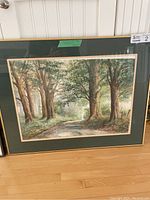 Full view of framed watercolour painting depicting a wooded forest path with large trees, green mat, and gold frame.