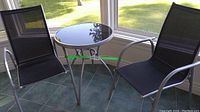 Two black mesh high back chairs with silver metal frames and a round glass metal frame table photographed indoors near a window.