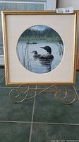 Framed acrylic painting showing geese swimming in a pond with a forest backdrop, with a gold frame and off-white matting in a circular window.