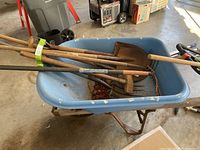Blue wheelbarrow containing multiple garden hand tools including shovels, rake, garden fork, and wooden mallet.