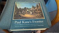 Front cover of the hardcover book titled 'Paul Kane's Frontier' showing a painting depicting Indigenous village life with tipis and people on the cover.