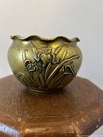 Front view showing embossed iris floral design and scalloped rim of brass bowl on wooden surface.