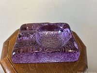 Square purple glass ashtray showing intricate internal texture detail, placed on wooden surface.