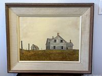 Front view of oil painting depicting rural house and smaller building with grassy field and neutral sky, framed with cream mat and wooden frame.