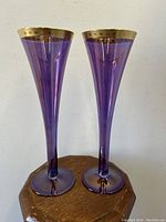 Front and side view of both royal purple iridescent art glass fluted vases with gold etched rims on a wooden octagonal surface against a plain wall