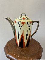 Front angle view of porcelain pot showing artistic geometric starburst red and black pattern on cream base with metallic silver handle and spout.