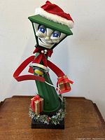 Front angle of the green lamp post figure with face, Santa hat, red bow tie, holding gifts, showing detailed facial expression and decorations.