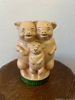 Front view of the Three Little Pigs cast iron still bank showing three pigs standing on green base, beige enamelled finish.