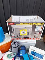 Photo showing Coleman portable electric cooler in original box, MEC blanket rolled up, MEC sleeping pillow in blue bag, Feja plastic dishes box, and some blue camping fabric items.