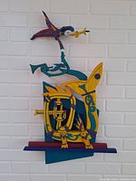Photo of colorful carved wood wall hanging mounted on white brick wall showing an abstract figure and fish-like shape with musical elements