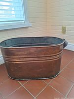 Photo showing full side view of the oval copper bin on a tiled floor near a window, showing copper color and missing handle.