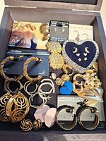 Photo showing an assortment of gold-tone hoops, clip-ons, and stud earrings including blue accent pieces inside a box.