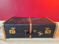 Closed view of Cunard vintage trunk showing black exterior, leather handle, brass latches, wood reinforcements and Cunard emblem.
