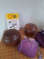 Wide photo showing wooden Go board with 19x19 grid, two round wooden containers, purple drawstring bags, and a strategy book.