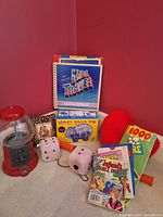Photo showing red bubble gum machine, several kids books including Kids Travel and Laugh Double Digest, Money Savvy Pig box, plush dice and red heart pillow arranged on the floor against a red wall.