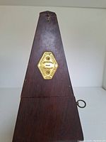Full front view of a wooden pyramid style metronome with a brass hexagonal plate centered on the front and visible winding key on the side.