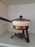 Side view of the mid century fondue pot with copper base, white porcelain dish, black steel stand, lid, and black handle.