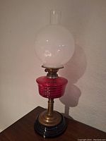 Full view of oil lamp showing red glass base, brass stem, and white glass globe chimney on black base.
