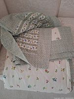 Folded quilt showing green and white small gingham checks, floral strips, and embroidered flower section.