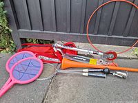 Silver Razor foldable scooter with carry bag, two paddle ball rackets, badminton racket, baseball bat, baseball glove, and jump rope arranged on the ground against a wooden backdrop