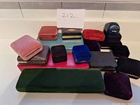A group of 15 small empty jewelry boxes in various velvet and leatherette finishes, different colors and shapes shown on a white counter.
