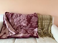 Two vintage throw blankets folded over a white couch, one burgundy with floral pattern and one beige with check pattern and fringe.