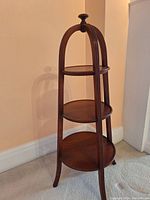 Full view of the wooden three-tier cake stand showing the tiers, legs, and arching supports against a plain wall and carpeted floor.