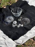 Crystal bowl, etched plates, cut glass spoon rests laid on black cloth outdoors, showing reflective qualities and clear glass patterns.