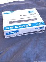Photo of Samsung DVD-E380 DVD Player retail box showing front side with branding, model, and features.