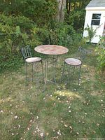 Full view of the outdoor high-top café table with two matching chairs on grass near bushes and a shed.